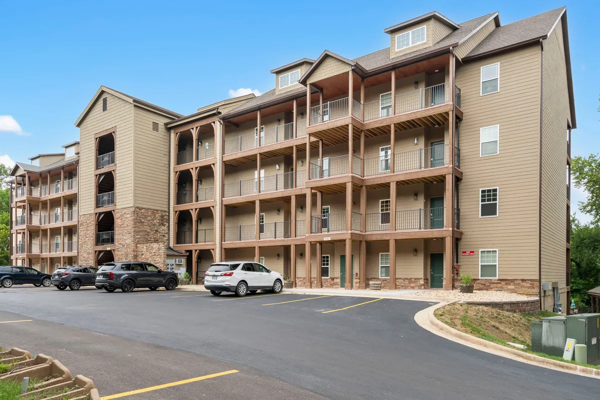 An inviting view of the building's stone facade, with balconies overlooking the green surroundings and a spacious parking area. The scene suggests moments of relaxation on private balconies, watching sunsets, and the ease of returning home after outdoor excursions.