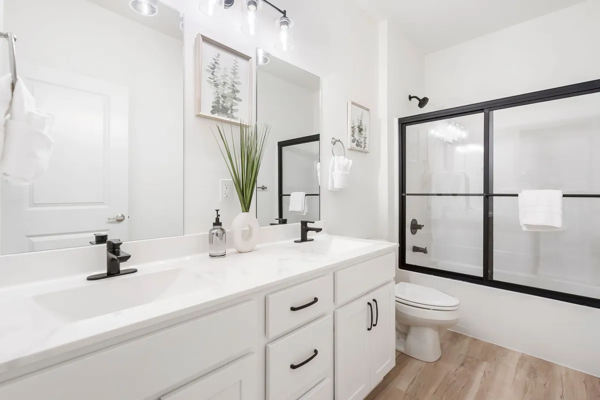 A spacious, bright bathroom with a double sink vanity, large mirrors, a glass shower enclosure, and a decorative plant. The clean, modern design feels inviting and perfect for morning routines or unwinding after a long day.