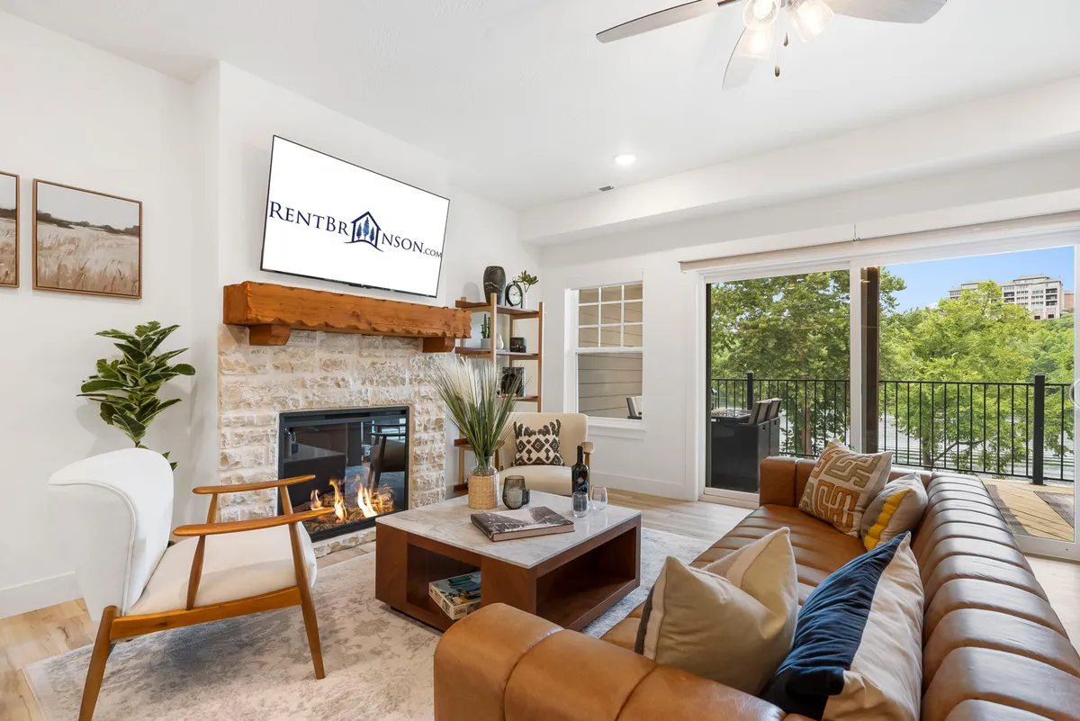 The image showcases a cozy living area with a plush sofa facing a warm fireplace, a large window framing scenic views, and a wall-mounted TV. The inviting ambiance suggests a perfect spot for relaxing evenings, whether by the fire or gazing out over Lake Taneycomo.