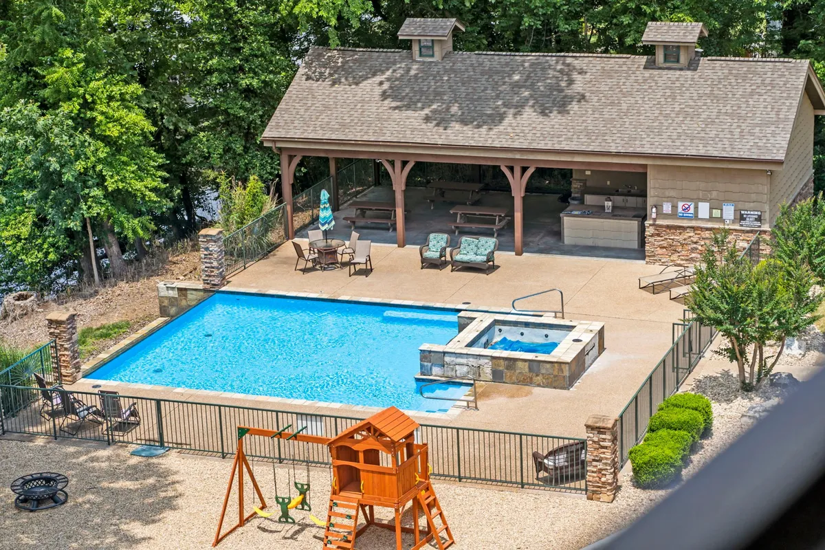 An exterior view of a resort-style swimming pool surrounded by lounge chairs, a hot tub, and a playset. The scene is lively, with children playing and families relaxing by the water. The covered patio area offers a comfortable spot for gatherings and barbecues.