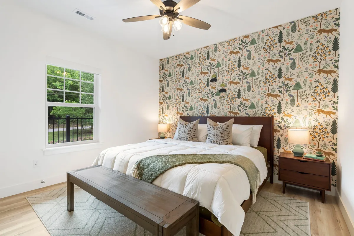 A tranquil King bedroom featuring a comfortable bed with decorative pillows, a feature wall with nature-themed wallpaper, wooden bench at the foot of the bed, and nightstands with warm lamp lighting. Soft natural light filters in, creating a soothing and inviting atmosphere ideal for relaxation.