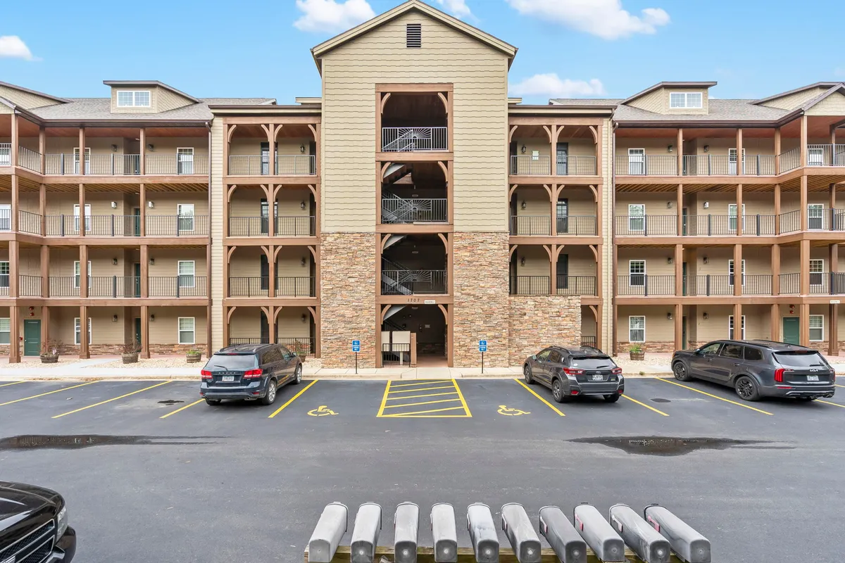This image displays the three-story building at Briarwood Shores, featuring stone and wooden accents, with a parking area and a staircase leading to the upper floors. The scene is bright and welcoming, showcasing the property's curb appeal and communal atmosphere.