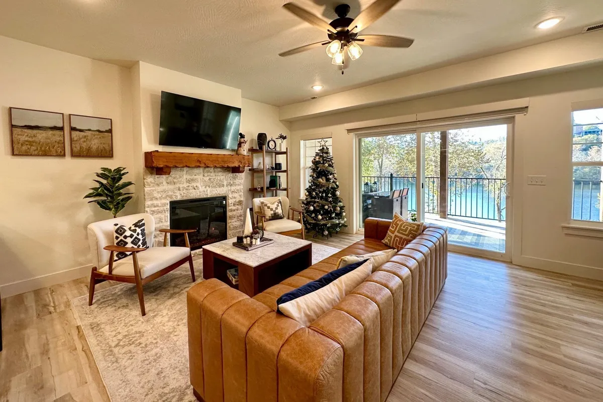 A spacious living room with a leather sectional couch, fireplace with a TV above, festive holiday décor, and large windows providing tranquil views. Shelves and a Christmas tree enhance the warm, welcoming atmosphere.